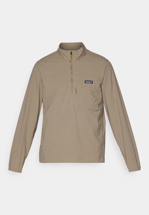 Beige hosszú ujjú pulóver fél cipzáros gallérral, elülső zsebbel és zöld cipzárhúzóval. Könnyű anyag, sima tapintással.