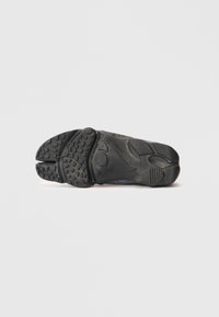 Fekete sneaker talp, amely textúrált gumiból készült, kiemelkedő lépésmintákkal, szellőzőnyílásokkal és a szélén domborított márkajelzéssel.