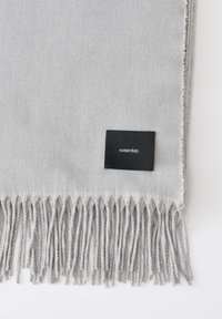 Grijze sjaal van zacht materiaal, met franjes aan de randen en een zwart label met witte tekst. Eenvoudig, strak ontwerp.