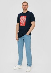 Marineblauw t-shirt met een rood-wit geometrisch grafisch ontwerp, gecombineerd met lichtblauwe rechte jeans en witte sneakers.