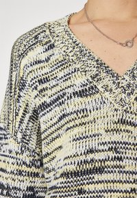 Pull en tricot avec un décolleté en V, motif zigzag jaune, noir et blanc, en tissu texturé et avec des bords ribblés.