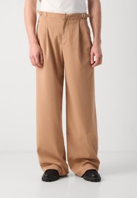 Pantalones beige de pierna ancha con una textura suave, que presentan un frente plano, trabillas para cinturón y detalles metálicos en la cintura. Estilizados con zapatos negros.