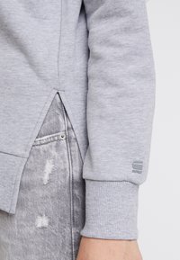 Grå sweatshirt med ribbade ärmslut, sidoklyftor i nederkant och en liten logopatch på ärmen. Strukturerad tyg med mjuk finish.