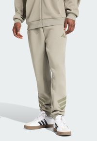 Lichtgrijze joggingbroek van een zachte stof, met olijfgroene drie-strepen accenten bij de enkels, gecombineerd met witte sneakers.