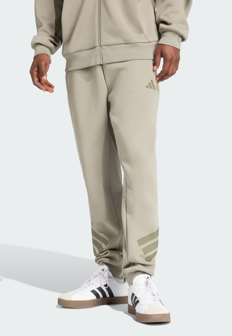 Lichtgrijze joggingbroek van een zachte stof, met olijfgroene drie-strepen accenten bij de enkels, gecombineerd met witte sneakers.