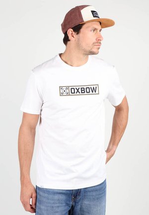 T-shirt en coton blanc avec le logo "OXBOW" en noir et un design de boîte olive. Porté avec une casquette brune et crème arborant un logo similaire. Jeans bleus.