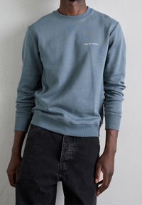 Hombre vestido con un sudadera de manga larga azul claro con el texto "Tiger of Sweden" en el pecho y jeans negros, frente a una pared lisa.