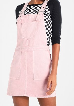 Robe salopette en denim rose avec deux poches avant et des bretelles réglables. Portée sur un haut à manches longues à carreaux noir et blanc.