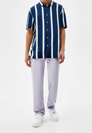 Camicia a righe blu navy e bianche a maniche corte, abbottonata, abbinata a pantaloni viola chiaro e sneakers bianche. Tessuto morbido, vestibilità rilassata, design casual.