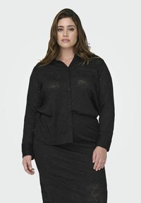 Chemise noire à manches longues avec un motif rose texturé. Le tissu semble léger, légèrement transparent et possède une coupe décontractée.