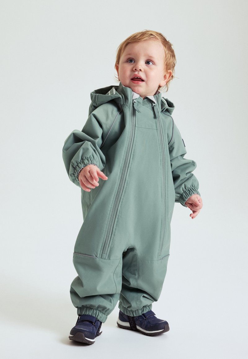 Peutertje staat in een te grote groene waterdichte sneeuwpak met twee voorzichtige ritsen en marineblauwe sneakers tegen een effen achtergrond.