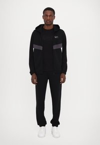 Black zip-up hoodie with gray stripe accents, paired with a black T-shirt and matching black sweatpants, completed with white sneakers.
