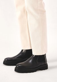 Botas de tobillo de cuero negro con punta redonda, paneles laterales de elastano y suelas de goma gruesas, combinadas con pantalones de color crema.