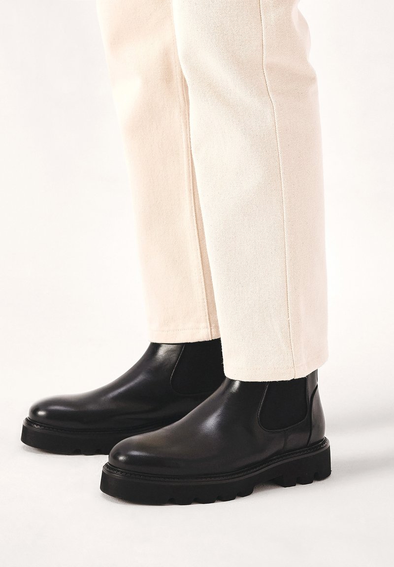 Botas de tobillo de cuero negro con punta redonda, paneles laterales de elastano y suelas de goma gruesas, combinadas con pantalones de color crema.
