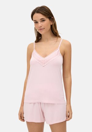 Joven mujer con camisola de encaje rosa claro y pantalones cortos a juego, de pie contra un fondo claro y sencillo.