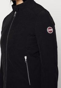 Černá bunda s vysokým límcem a předním zipem. Má boční kapsu na zip se stříbrným zipem a kulatou logo nášivku na rukávu.