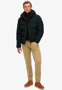 Puchowa ciemnozielona kurtka z kapturem na brązowym golfie, w połączeniu z beżowymi spodniami i brązowymi sznurowanymi butami, stojąca na neutralnym tle.