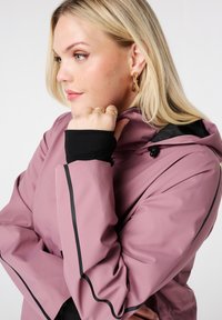 Blonde vrouw met een mauve jasje met zwarte accenten en een capuchon, die haar kin rustend op haar hand steunt, versierd met gouden ringen en cascade oorknopjes.