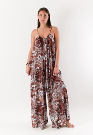 Donna in piedi che indossa una tuta larga marrone con stampa floreale, con spalline sottili e gambe larghe, su uno sfondo bianco semplice.