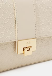 Béžová kabelka s vyraženým kruhovým vzorem, hladkým koženým páskem a zlatým zapínáním s vyrytým logem.