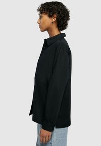 Zwarte overhemd met lange mouwen, één borstzak en een klassieke kraag. Draagt lichtblauwe jeans, weergegeven tegen een neutrale achtergrond.
