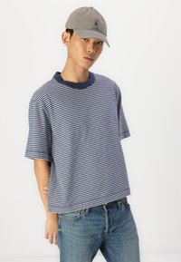 Navy en witte gestreepte T-shirt met korte mouwen en een ribbelkraag in navy. Gemaakt met blauwe jeans en een lichtgrijze baseballcap.