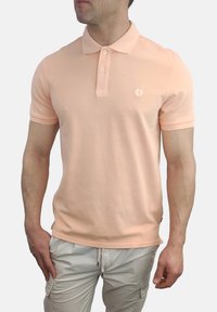 Camisa polo de melocotón con cuello clásico, mangas cortas y un logotipo sutil en el pecho. La tela parece suave y liviana.