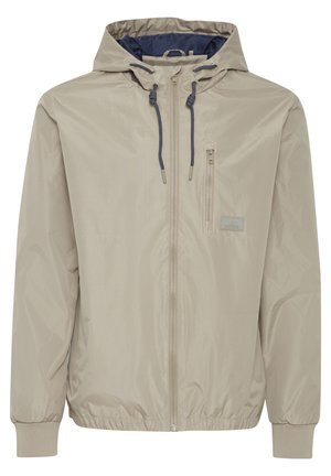 Giacca zip-up color beige chiaro con cappuccio, polsini elastici e tasca sul petto con un'etichetta logo rettangolare. Tessuto liscio e resistente all'acqua.