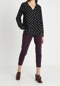 Blouse noire à manches longues avec un motif d'arbre blanc, design à col en V ; associée à un pantalon ajusté violet et des chaussures à talons brillantes marron.