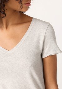 Žena s kučeravými vlasmi oblečená v svetlošedej tričku s výstrihom do V a krátkymi rukávmi, zobrazená od krku po hornú časť paže na jednoduchom pozadí.