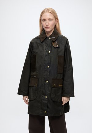 Frau mit langen blonden Haaren trägt einen dunkelgrünen Barbour-Mantel mit braunem Cordkragen und Taschen, steht vor einem schlichten weißen Hintergrund.