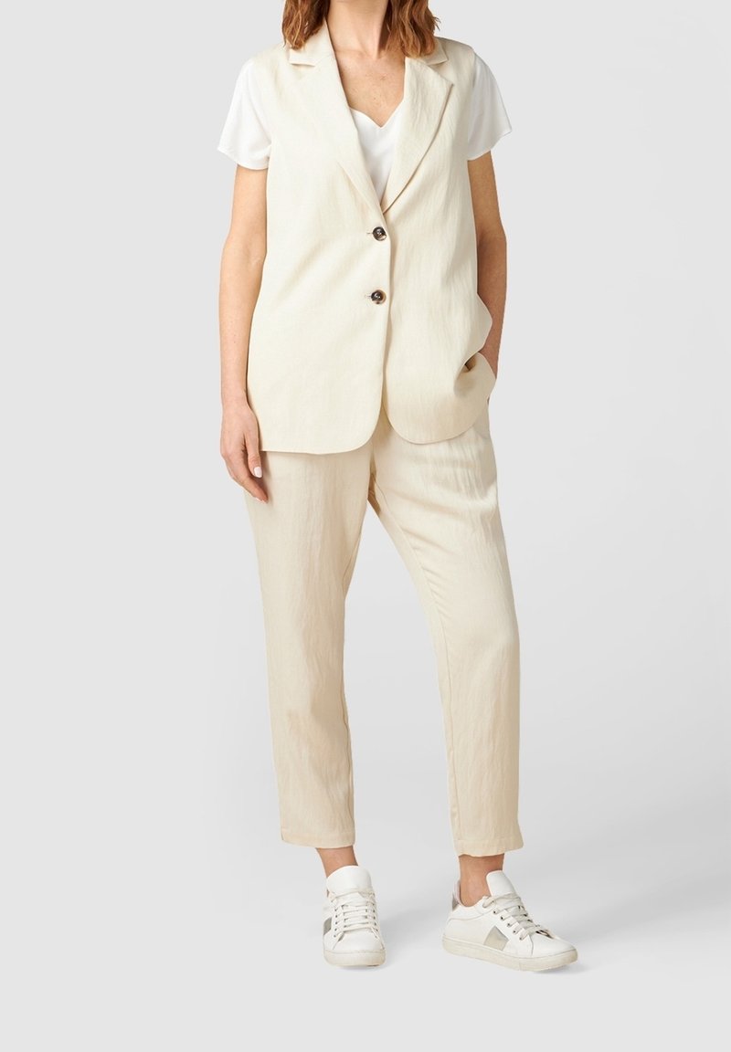 Krämfärgad linneväst med urklippt krage och två knappar, i kombination med matchande smalbyxor och vita sneakers med grå detaljer.
