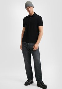 Uomo che indossa un berretto grigio a maglia, una polo nera a maniche corte, jeans grigio scuro e scarpe nere con suola spessa, in piedi contro uno sfondo chiaro uniforme.
