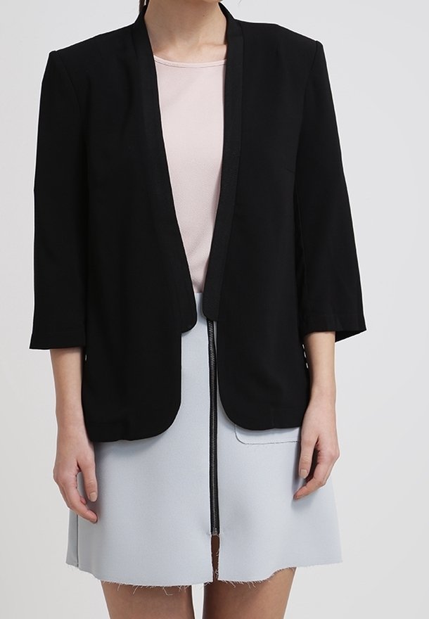 Kobieta w czarnym żakiecie bez zapięcia, nosząca jasnoróżową bluzkę i jasnoszary spódnicę z zamkiem z przodu oraz detalami kieszeni, stojąca na tle jednolitego tła.