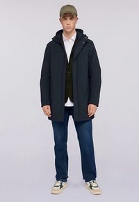 Tmavě modrá nepromokavá bunda s kapucí, přes černou vestu a bílou košili, doplněná modrými džínami a teniskami se zelenými prvky.