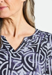 Lila Bluse mit V-Ausschnitt, versehen mit einem weißen abstrakten Muster. Der Stoff hat eine glatte, glänzende Textur und eine subtile Raffung an den Schultern.