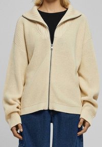Béžový svetr na zip s vysokým žebrovaným límcem, širokými rukávy a pletenou texturou. Má volný střih a kontrastní černý zip.