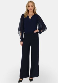 Granatowy kombinezon z przezroczystymi długimi rękawami, dekoltem w kształcie litery V i szerokimi nogawkami. Akcentowany dekoracyjnym łańcuszkiem przy dekolcie. Teksturowana tkanina.