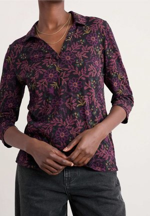 Donker bloemenpatroon shirt met lange mouwen met paarse en groene bloemen, gedragen met een donkere spijkerbroek en gelaagde gouden kettingkettingen.