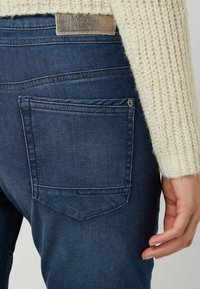 Dark blue denim jeans with a rectangular back pocket, visible stitching, and a silver button accent on top. Fabric displays subtle texture.