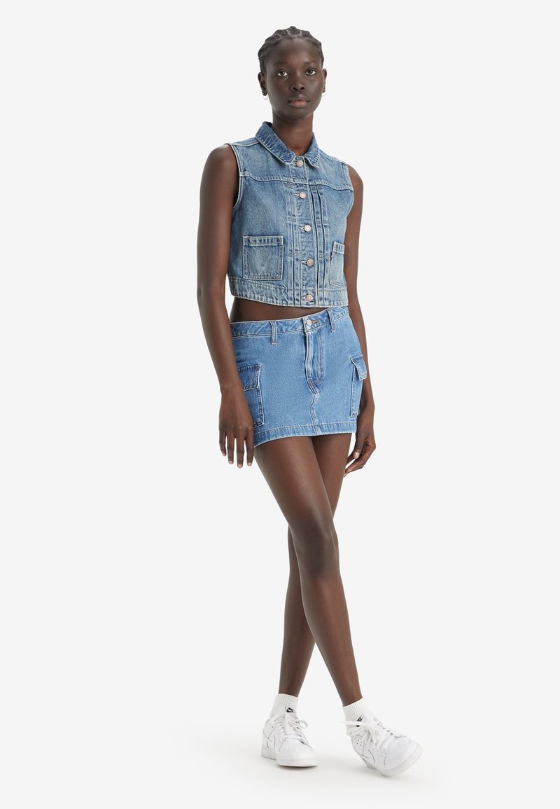 Gilet in denim corto con colletto, bottoni frontali e due tasche, abbinato a una gonna mini in denim coordinata con tasche. Entrambi gli articoli sono in azzurro chiaro.