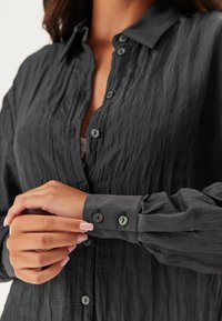 Camicia button-up nera in tessuto stropicciato con colletto; presenta bottoni arrotondati, dettagli sui polsini e una vestibilità rilassata. Superficie testurizzata.