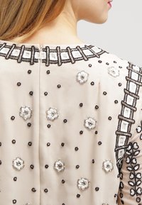 Dos et épaule supérieurs d'une femme vêtue d'un habit beige orné de broderies florales et géométriques en perles noires et blanches.