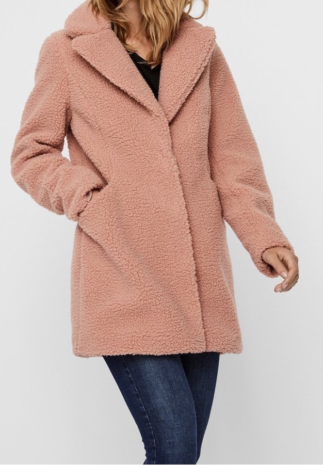Femme portant un manteau teddy rose clair à large col et poches latérales, associé à un jean bleu foncé sur un fond uni.