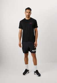 Melna īsroku T-krekls ar rozā logotipu, kombinēts ar melniem šortiem ar baltu "NIKE" uzrakstu, baltām sporta zeķēm un melniem apaviem.