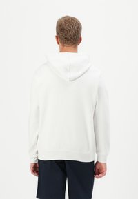 Hvit hettegenser med snorhette, ribbestrikkede mansjetter og kant. Glatt tekstur, avslappet passform, vist fra baksiden mot en ensfarget bakgrunn.