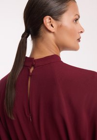 Bordowa sukienka z wysokim kołnierzem, detalem w kształcie dziurki na plecach oraz niewidocznym zamkiem. Gładki materiał elegancko układa się na sylwetce.