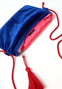 Sacca di velluto blu con interno rosa, zip dorata e nappina rossa. Presenta un cordoncino intrecciato rosso per la chiusura e il trasporto.