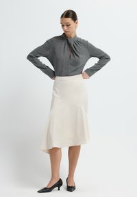 Grijze blouse met lange mouwen en een sleutelgatdecolleté, gecombineerd met een crèmekleurige midirok met een asymmetrische zoom en subtiele draping. Zwarte hakken.