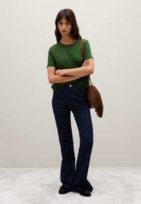 Grünes T-Shirt mit kurzen Ärmeln, dunkelblaue Schlagjeans und braune Fransenhandtasche. Das Model steht mit verschränkten Armen auf einer hellen, strukturierten Oberfläche.